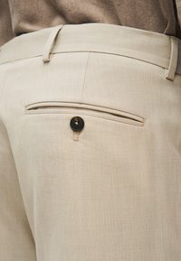 Κοντινό πλάνο μπεζ παντελονιού φόρμας που δείχνει μια πίσω τσέπη με μαύρο κουμπί και θηλιές για ζώνη, φορεμένο με καφέ τοπ.