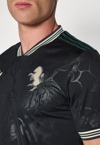 Maillot de sport noir à col en V, avec un col rayé vert et blanc. Motif floral en relief sur le tissu, avec un emblème contrastant sur la poitrine.