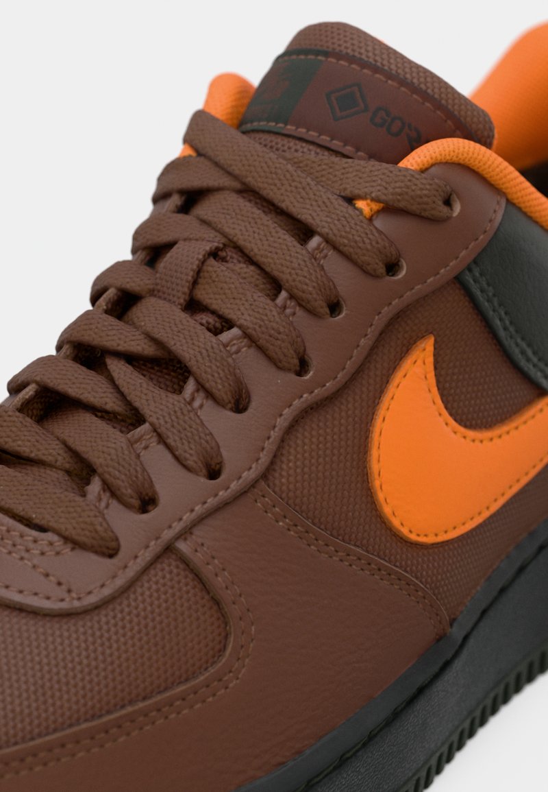 Baskets de sport marron et orange avec des panneaux texturés et lisses, des lacets marron et un swoosh Nike orange sur le côté.