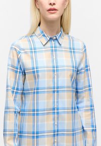 Frau, die ein langärmliges Hemd mit einem karierten Muster in Blau, Beige und Weiß trägt, steht vor einem schlichten weißen Hintergrund.