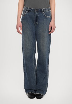 Jeans de mezclilla azul de pierna ancha con un acabado desgastado, que presentan herrajes plateados, diseño de cinco bolsillos y corte recto, llevados sobre zapatos negros.