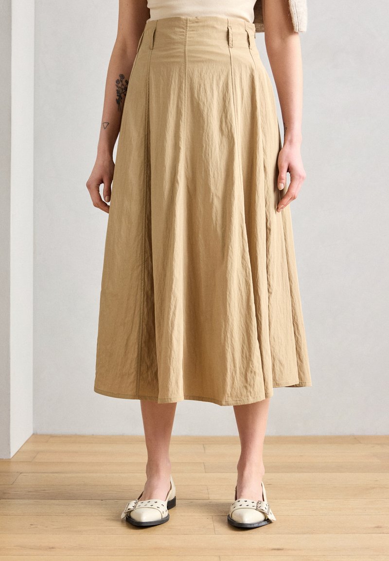 Jupe beige arrivant aux genoux en tissu froissé. Elle présente une taille haute, des plis et des coutures latérales. Associée à des chaussures blanches et un fond en bois.