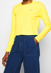 Långärmad gul bomullströja med rund halsringning och FILA-logotyp. Matchad med höga mörkblå denimbyxor.