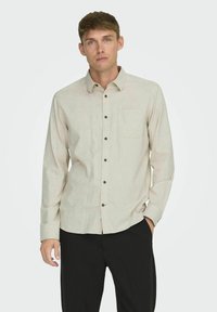 Camisa de manga larga color beige claro con cuello abotonado, bolsillo en el pecho y botones marrones, fabricada con un material texturizado.