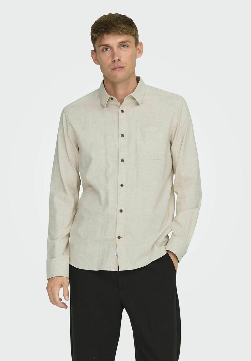 Camisa de manga larga color beige claro con cuello abotonado, bolsillo en el pecho y botones marrones, fabricada con un material texturizado.