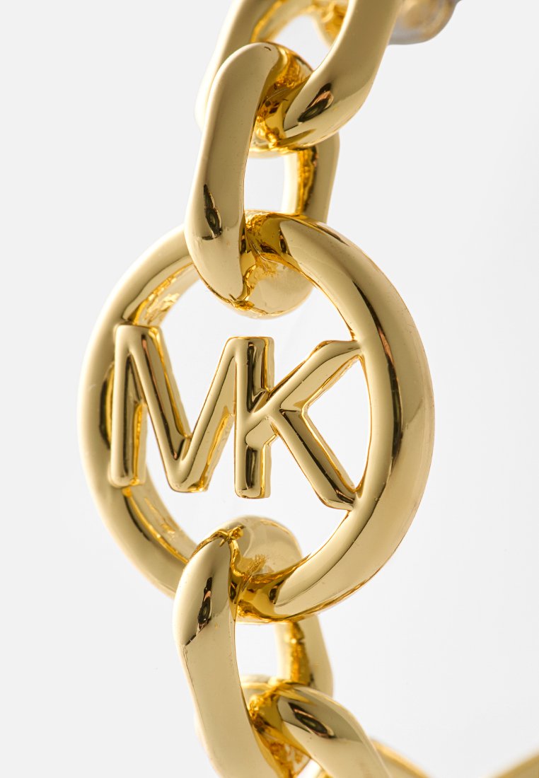 Close-up van een glanzende gouden ketting met een ronde hanger met het verhoogde "MK"-logo in het midden op een witte achtergrond.