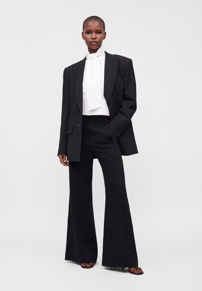 Personne portant un blazer noir oversize, pantalon noir à jambes larges, blouse blanche à col montant avec un détail devant, debout avec une main dans la poche.