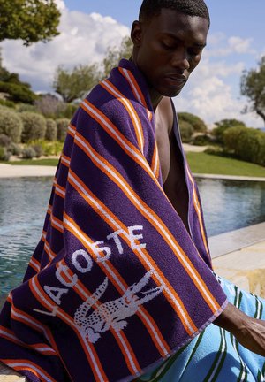 Man sitting by pool wrapped in purple, orange, and white striped Lacoste towel on a sunny day with green shrubs and blue sky.
