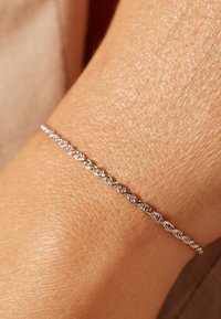 Bracelet en argent tordu porté sur une surface de peau texturée, présentant un design délicat et entrelacé avec une finition lisse.