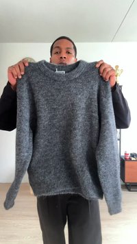 Suéter gris de punto con cuello redondo, ajuste holgado y superficie texturizada. Presenta mangas largas y un dobladillo trasero ligeramente más largo.