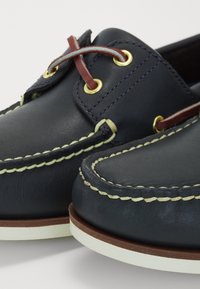 Chaussures bateau en cuir marine avec coutures crème, oeillets dorés et accents marron. Texture lisse avec un design à bout rond et semelle plate blanche.