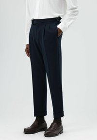 Pantalon plissé bleu marine avec taille ajustable, ourlets retroussés et poches latérales, associé à des mocassins marron foncé et une chemise blanche à manches longues.