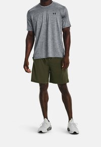 Muž v šedém tričku s krátkými rukávy, olivově zelených šortkách a bílých běžeckých botách stojí na bílém pozadí.