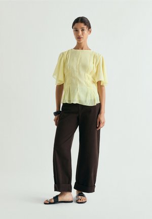 Mujer de pie con una blusa amarilla clara con mangas con volantes, pantalones anchos oscuros con puños en los tobillos, sandalias negras y una pulsera negra.