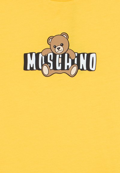 Καφέ αρκουδάκι κρατάει στιλιζαρισμένα ασπρόμαυρα γράμματα που σχηματίζουν το όνομα μιας μάρκας σε κίτρινο φόντο.