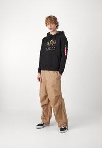 Felpa nera con logo dorato, tasca frontale e tasca laterale sulla manica; pantaloni ampi beige con una texture stropicciata; sneakers in bianco e nero.