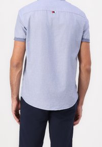 Camicia a maniche corte con bottoni in tessuto azzurro chiaro con una sottile trama. Orlo arrotondato e una piccola etichetta con logo sul retro del collo.