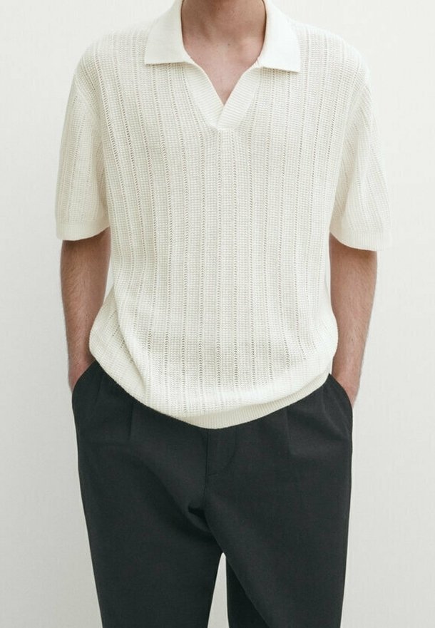 Homme portant un polo en maille blanche à manches courtes, rentré dans un pantalon noir, les mains dans les poches, sur un fond uni.
