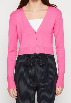 Femme portant un cardigan rose vif boutonné sur un haut blanc avec un pantalon noir taille haute à fines rayures, noué à la taille.