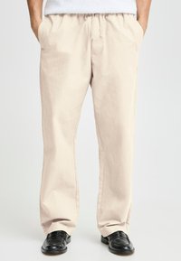 Beige cotton trousers with an elastic waistband, side pockets, relaxed fit, and slight texture, paired with black leather shoes.