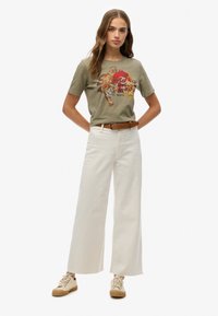 Camiseta verde oliva con un gráfico de tigre, combinada con pantalones anchos blancos. Zapatillas beige con acentos marrones completan el atuendo.