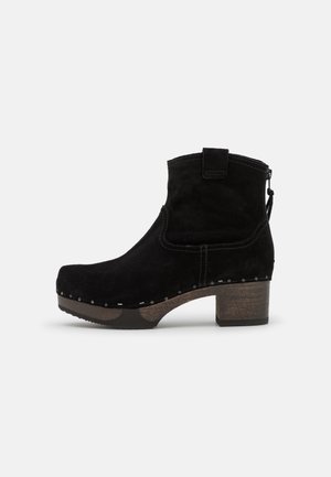 Botín de ante negro con tacón de madera apilada, cremallera lateral y tachuelas metálicas decorativas a lo largo del borde de la suela.