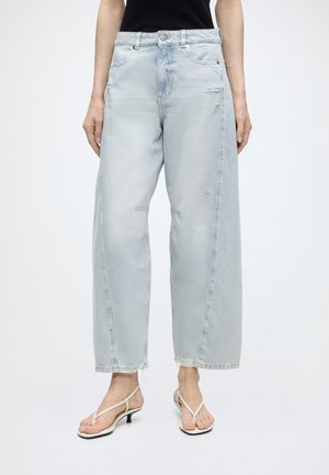 Personne portant un jean bleu clair large et usé avec des ourlets effilochés, associé à des sandales blanches à brides et talons bas, et un haut noir.