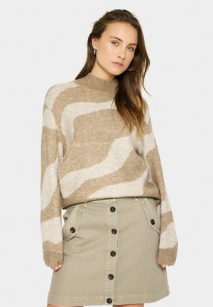 Maglione beige a maglia con motivo ondulato bianco, collo alto e maniche lunghe, abbinato a una gonna button-up beige chiaro con tasche laterali.