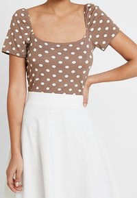 Top ajustado marrón con lunares, mangas cortas abullonadas y escote cuadrado, combinado con una falda blanca de corte en A. Textura simple y suave del tejido.