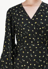 Black wrap dress featuring a floral pattern with yellow flowers and green leaves, long sleeves, and a tied waist detail. Soft fabric texture.