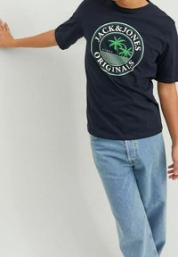 Mörkblå t-shirt med ett cirkulärt grafiskt motiv med palmer och texten "JACK&JONES ORIGINALS". Kombinerad med ljusblå jeans.