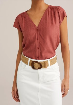 Kvinde iført en rustfarvet, tekstureret V-hals skjorte med knapper, stukket ned i et hvidt skørt med et vævet bælte med en stor skildpaddepanser spænde.