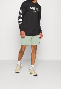 Czarna koszula z długim rękawem z białymi logo, zestawiona z jasnoszarymi szortami i beżowymi butami sportowymi z zielonymi akcentami.