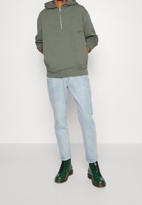 Sudadera verde con cremallera y bolsillo tipo canguro, combinada con jeans azul claro y botas de cuero verde oscuro con cordones. Un conjunto simple y casual.