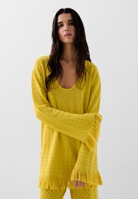Geel gebreid vest met een diepe halslijn, franjes aan de onderkant en gestructureerde patronen. Vergezeld van bijpassende gele leggings met een textuur.