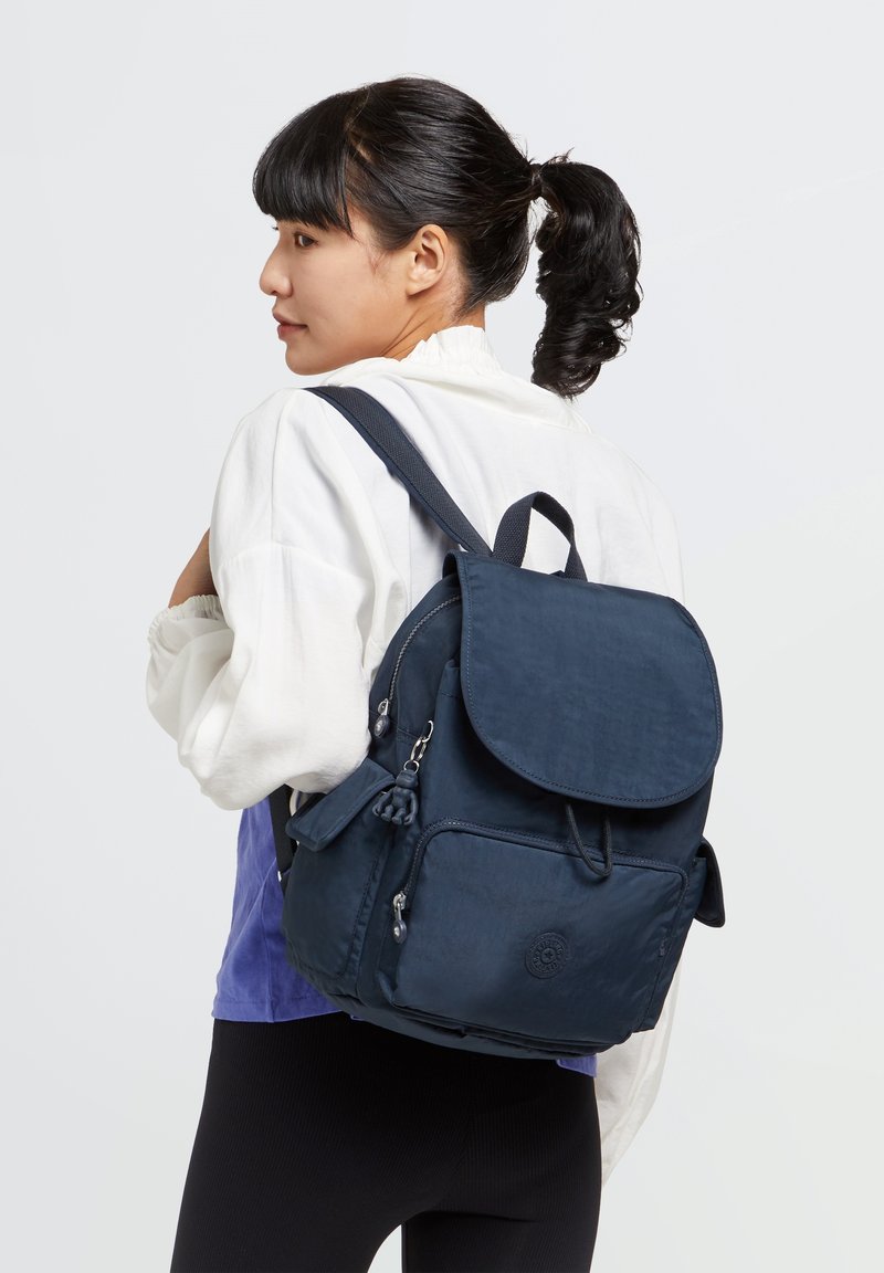 Mochila de tela azul marino con solapa, múltiples bolsillos con cremallera, correas ajustables y un detalle de logo circular en la parte frontal.