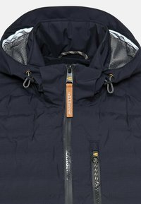 Navy puffer jacket with a high collar, contrasting inner lining, front zipper, and drawstring hood. Features logo tags and textured fabric.