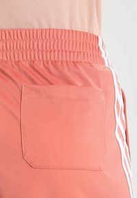 Korálové sportovní šortky s elastickým pasem, bílými pruhy po stranách a obdélníkovou zadní kapsou; hladká textura a lehká látka.