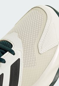 Sneaker de sport blanche avec des panneaux en maille texturée, des superpositions synthétiques lisses et des lacets vert foncé. Comprend des accents noirs sur le côté.