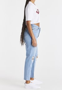 Weißes, gekürztes T-Shirt mit roter Aufschrift, kombiniert mit hellblauen, zerrissenen Jeans und weißen Turnschuhen mit schwarzen Akzenten. Seitenansicht.