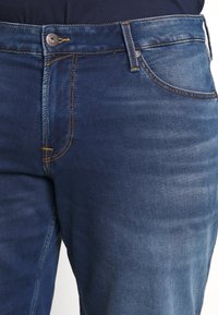 Detailní záběr na modré džínové kalhoty s oranžovým šitím, kovovým knoflíkem a přední kapsou, nošené s tmavým topem.