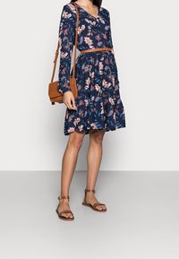 Robe florale bleue à manches longues, décolleté en V et taille cintrée. Associée à des sandales brunes et un sac à bandoulière assorti.