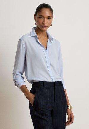 Vrouw die een lichtblauw button-up overhemd met opgerolde mouwen en een donker gestreepte broek draagt, staand met één hand in de zak.