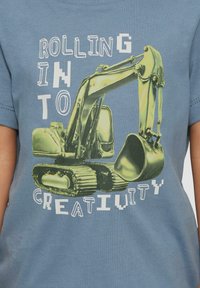 Lichtblauwe t-shirt met een afbeelding van een gele graafmachine en de tekst "ROLLING INTO CREATIVITY" in vetgedrukte, gevarieerde lettertypes.