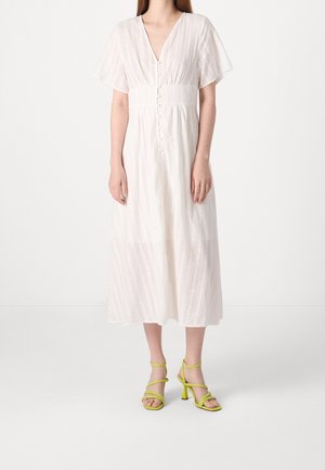 Frau trägt ein weißes Midikleid mit kurzen Flatterärmeln und Knopfdetails vorne, kombiniert mit limettengrünen Riemchen-Sandalen mit Absatz.