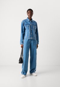 Giubbotto in denim blu con dettagli strappati, abbinato a jeans a gamba larga con orli sfrangiati. La modella tiene in mano una borsa nera e indossa sneakers bianche.