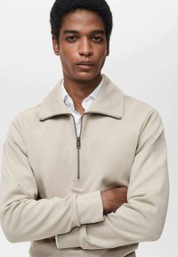 Felpa con zip di colore beige chiaro, dotata di un colletto alto, con maniche lunghe e una texture liscia. Nessun motivo o accento visibile.