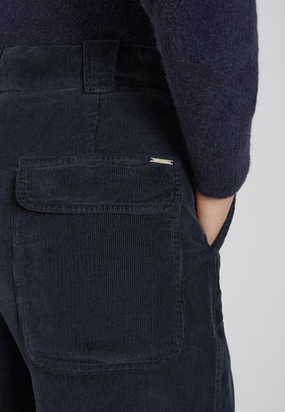 Donkerblauwe corduroy broeken met een gestructureerde afwerking, voorzien van een enkele achterzak en een metalen merkteken voor detail.