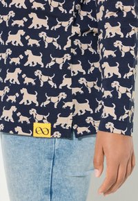 Marineblaues Hemd mit beigem Hundemuster, mit kurzen Ärmeln und einem Seitenschild mit gelbem Quadrat und Unendlichkeitssymbol. Weicher Stoff und entspannte Passform.