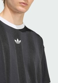 Melnas svītrains T-krekls ar baltu apkakli un Adidas uzrakstu. Tekstūra izskatās gluda, ar nedaudz spīdīgu apdari.